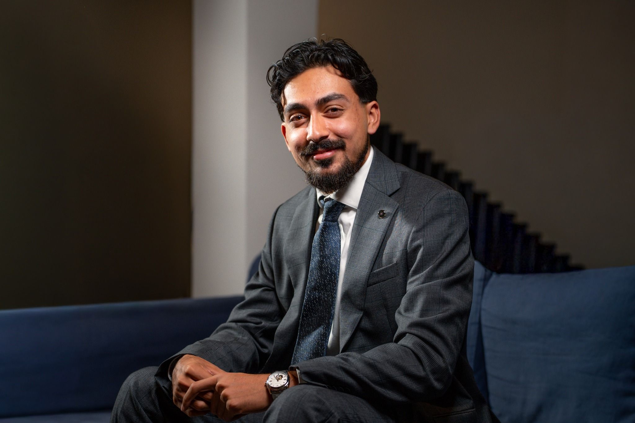 Siraaj Shah seated on a blue sofa, smiling, in a grey suit